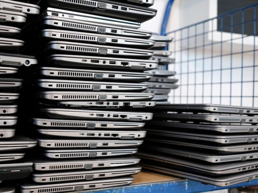 Several stacks of closed laptop computers are arranged on shelves, showcasing their edges with visible ports and vents—ready for processing as part of a laptop buyback program.