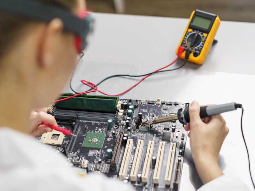 Person soldering a component on a computer motherboard with a soldering iron; a digital multimeter for electrical testing is positioned nearby on the work surface.
