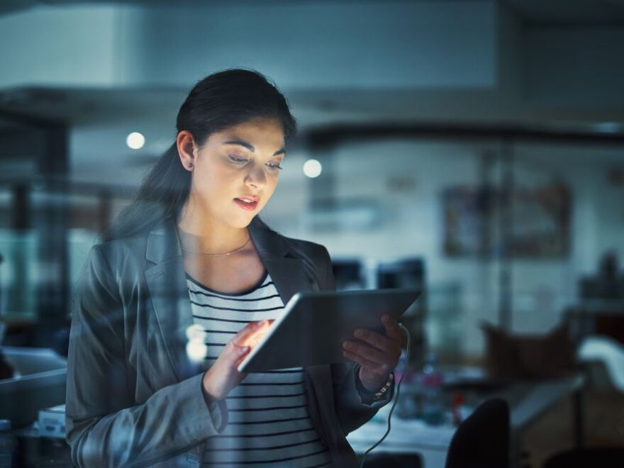 A woman in business attire stands indoors, using a tablet device—an image reflecting the dynamic environment of tech recruitment Singapore, with modern office equipment visible in the background.