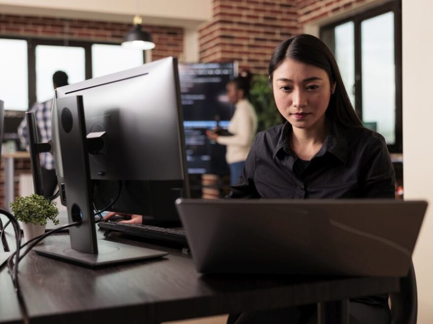 Woman working at a desk with a laptop and desktop computer in a modern office, where she managed Cisco Meraki network setups for small Singapore offices, with two coworkers and large windows in the background.