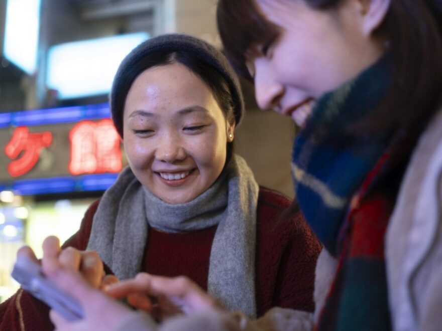 Two women wearing scarves and winter clothes smile while looking at a smartphone outdoors at night, with neon signs in the background, possibly exploring Tech-and-GO funded technology upgrades for charities across Singapore.