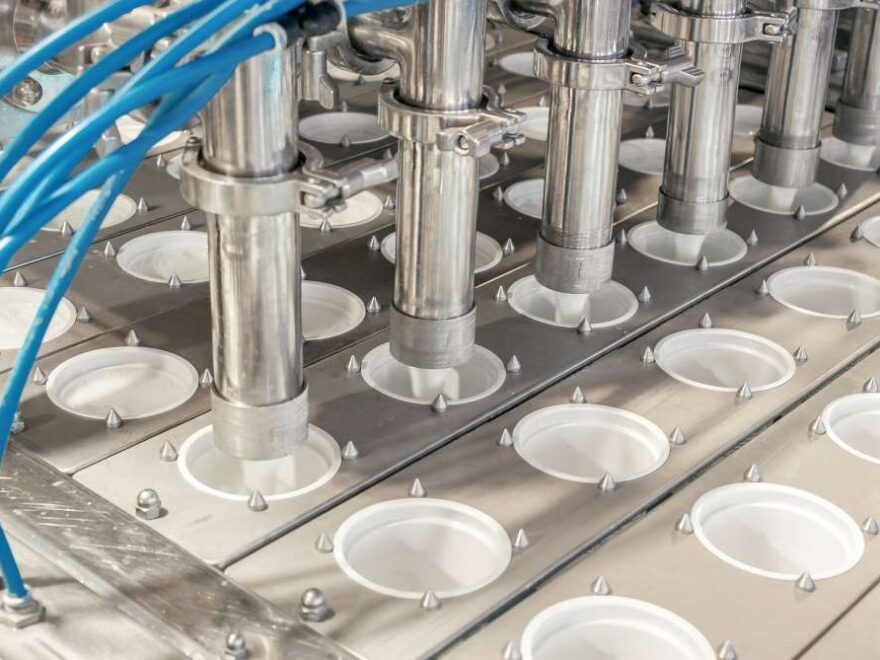 Automated filling machine dispensing liquid into multiple round containers arranged in rows in a factory setting.