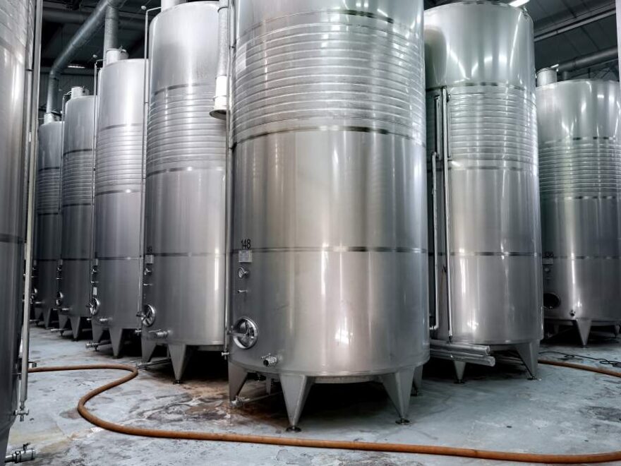 Rows of large stainless steel tanks in an industrial facility, with pipes and hoses on the concrete floor.